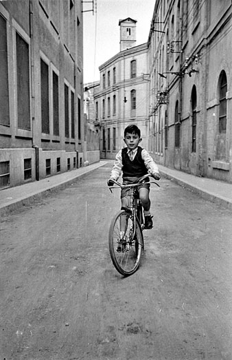 Retrat d'un nen amb bicicleta al carrer de la Sèquia. A banda i banda, la fàbrica Grober. Al fons, la torre de les Aigües. 1955