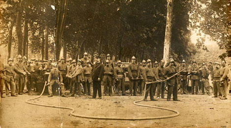 Retrat de grup de la Primera Companyia de Bombers de l'Ajuntament de Girona, amb els seus uniformes i recursos materials per lluitar contra el foc, a la Devesa. Possiblement es tracti d'una de les exhibicions que es portaven a terme durant les fires. 1900