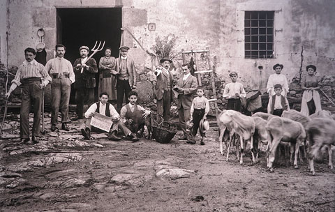 Grup de pagesos davant d'una masia. A l'esquerra, homes amb eines de conreu. A la dreta, dones i nens amb un ramat d'ovelles. Sense identificar. 1910