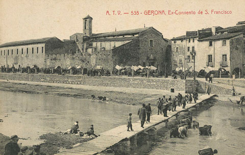 El convent de Sant Francesc de Paula, conegut com els Mínims, i la palanca d'en Vila o de l'Areny vistos de l'altre costat del riu. A lesquerra, paral·lel al riu, el passeig General Mendoza ple de carruatges durant un dia de mercat i a la dreta, la rambla Pi i Margall. S'observen bugaderes rentant roba al riu. 1905-1907