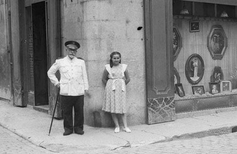 Façana i aparador de la galeria Foto Lux en l'encreuament dels carrers Ciutadans i Cort Reial. S'observa el Cap de la Policia Municipal, el senyor Romero i a Joana Pereferrer. Sobre la galeria hi ha el local de la Secció Femenina de la Falange. 1950-1955