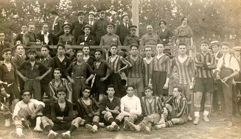 Partit de futbol entre la Unió Deportiva Girona i el Calella al camp de futbol de la Devesa, al Camp de Mart, en benefici dels soldats que lluitaven a l'Àfrica. Retrat de grup dels jugadors davant la tribuna d'autoritats civils i militars presents a l'encontre. S'identifica a Jesús Ballestero, president del club. Entre els jugadors, a d'alt d'esquerra a dreta: Viarnés, Padrosa, Bisbe, Viñas, Alvarez i Roca. Asseguts: Casteyó, Pep Gómez, Prats, Fernández i Rustey. 18/02/1921