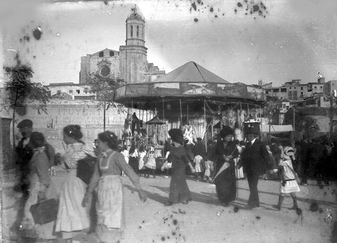 Atraccions de Fires a la plaça de la Independència. Al fons, un anunci d'un reconstituent pintat a la paret que tancava la plaça per la banda del riu, la part inferior de la qual encara conserva trets de la fortificació que donava a l'Onyar. El mur desparagué el 1914, amb la urbanització del passeig Canalejas. 1900
