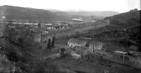 Vista del monestir de Sant Pere de Galligants i del tram de muralla que enfila cap a Montju&iuml;c. En primer terme, el carrer de Sant Daniel que mena cap a la vall. Al centre, adossat a la muralla, el tambor de Sant Daniel i el portal de Sant Pere. A la dreta, la torratxa de Santa Ll&uacute;cia i la muntanya de Montju&iuml;c amb el castell hom&ograve;nim. 1896-1911