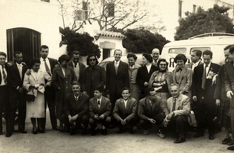 Celebració de la festa de l'Arcàngel Gabriel, patró dels radiofonistes. Retrat de grup dels treballadors de Radio Gerona EAJ38 i altres assistents davant d'un restaurant d'un lloc no identificat. S'identifiquen, ajupits, el primer a l'esquerra, Emilio Banda. Drets, la setena a la dreta, Francina Boris, el sisè, Miquel Vidal i el primer a la dreta, Jaume Teixidor. 1955